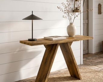 May include: A wooden console table with a V-shaped base, featuring a dark brown wood grain finish. On the tabletop sits a black metal lamp with a conical shade and a white ceramic vase containing thin, leafless branches.