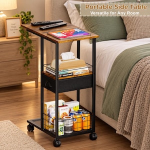 May include: A portable side table with a black metal frame and two wood-grain shelves. It features a fabric drawer, a bottom wire storage basket, and four caster wheels. The top surface holds two remote controls and a tablet.