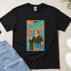 May include: A black short-sleeve t-shirt featuring a graphic print of a Tony Soprano action figure in its original packaging. The design includes the text The Sopranos and Tony Soprano over a colourful, swirling, psychedelic background pattern.