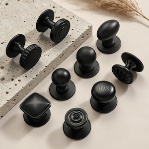 May include: A collection of nine matte black cabinet knobs in various shapes, including round, square, and fluted designs. One round knob features the letters HWS engraved on its surface. The knobs are arranged on a light-colored surface.