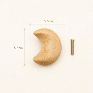 May include: A crescent moon-shaped wooden knob with a light tan wood grain finish. The knob measures 5.5 cm in width and 5.5 cm in height. A single metal screw with a threaded shaft and a flat head is positioned to the right of the wooden knob.