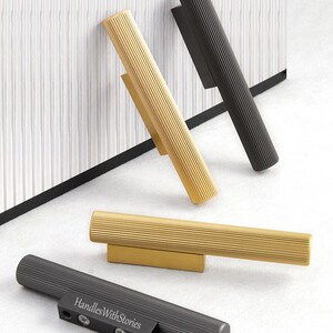 May include: Four cylindrical cabinet handles with a ribbed texture, featuring two in a gold finish and two in a matte black finish. One black handle displays the text "HandlesWithStories" on its rectangular mounting base with visible screws.