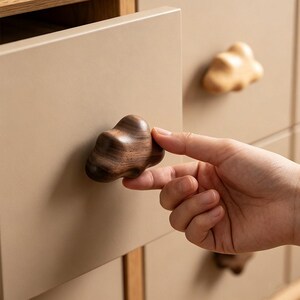 May include: A dark brown wooden drawer knob shaped like a cloud attached to a matte beige drawer front. The knob features a smooth, rounded surface with visible wood grain patterns. A second, lighter-coloured wooden cloud knob is visible on the drawer above.