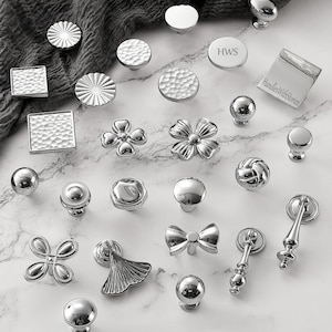 May include: A collection of silver-toned metal cabinet knobs and pulls in various shapes, including round, square, floral, and bow designs. Some pieces feature hammered textures, smooth polished finishes, or engraved text reading HWS and HandlesWithStories.