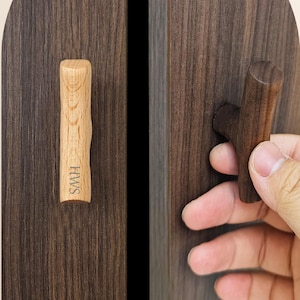 Puede incluir: Dos tiradores de armario de madera en forma de T montados sobre paneles de madera de color marrón oscuro. El tirador izquierdo es de madera clara con las letras HWS grabadas, mientras que el derecho es de madera oscura y lo sujeta una mano.