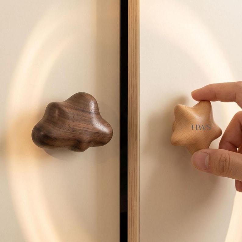 May include: Two wooden cabinet knobs attached to a light-coloured door. One knob is dark brown with a smooth, irregular cloud-like shape. The other knob is light tan, star-shaped, and features the letters HWS printed in a dark font on its surface.