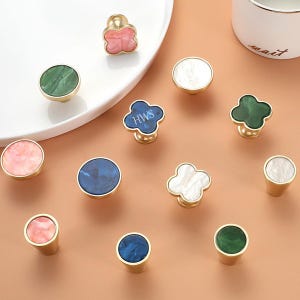 Puede incluir: Una colección de pomos para armarios con bases de metal dorado y tapas decorativas de diversas formas, incluyendo diseños circulares y de trébol con patrones de mármol en rosa, verde, blanco y azul, con un pomo azul que muestra el texto HWS.
