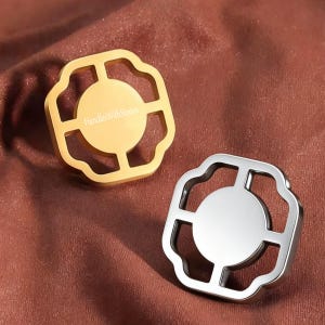 Può includere: Due pomelli per armadi in metallo con cornice quadrata dai bordi smerlati e centro circolare. Un pomello è color oro con il testo "HandlesWithStories" inciso al centro, l'altro è color argento con un centro circolare liscio e semplice.
