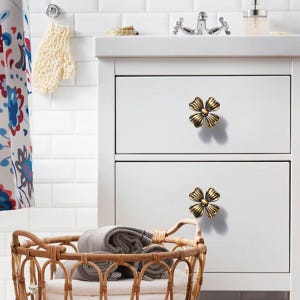 May include: Two antique brass-toned metal drawer knobs shaped like four-petaled flowers. Each knob features a central circular detail with radiating, textured petals, mounted on the front of two stacked white rectangular cabinet drawers.