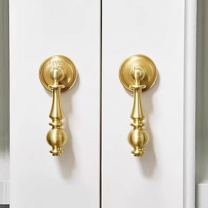 May include: Two matching brass drop pulls with a brushed finish, each featuring a circular backplate and a vertical, tiered handle. The left pull is engraved with the letters HWS on the top circular plate, mounted on white cabinet doors.