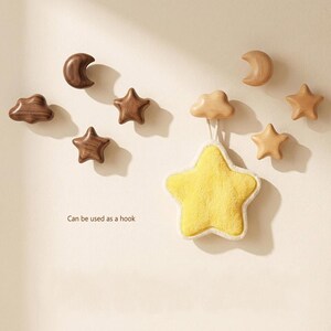 May include: A collection of wall-mounted wooden hooks shaped like moons, stars, and clouds in dark and light wood tones. One light-coloured cloud hook supports a plush yellow star-shaped item. The text "Can be used as a hook" is visible.