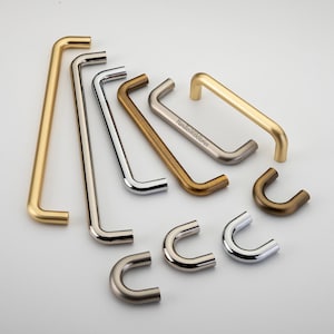 Puede incluir: Una colección de tiradores de metal para armarios en varios acabados, incluyendo oro cepillado, cromo pulido y bronce antiguo. El surtido presenta tiradores de barra largos y tiradores pequeños en forma de U, con el texto HandlesWithStories.
