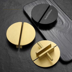 May include: Two brushed gold semi-circular cabinet pulls and one black semi-circular cabinet pull with the text HandlesWithStories printed on the surface. The gold pulls show a flat front and a rear mounting bracket with two protruding metal posts.
