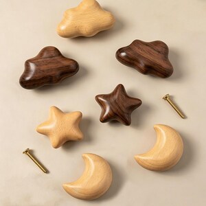 May include: A collection of wooden cabinet knobs in the shapes of clouds, stars, and moons, featuring both light-toned beech wood and dark-toned walnut wood finishes. Two metal mounting screws are included alongside the seven wooden shapes.