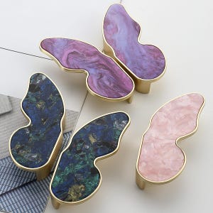Peut inclure: Quatre boutons de placard en forme de papillon avec des bords en métal doré. Les boutons présentent des inserts en résine marbrée de couleur violette, bleu profond avec des éclats verts et dorés, et rose clair. Silhouette irrégulière.