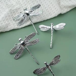 May include: Four silver-toned dragonfly-shaped cabinet knobs with intricate wing detailing. One of the dragonfly knobs features the letters HWS embossed on the upper wing. Each knob has a slender body and extended wings, designed for furniture hardware.