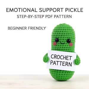 Könnte beinhalten: Eine leuchtend grüne gehäkelte Gurkenfigur mit schwarzen Sicherheitsaugen, rosa Wangen und einem kleinen Lächeln. Die Figur hält ein weißes Schild mit der Aufschrift CROCHET PATTERN. Der Text darüber lautet EMOTIONAL SUPPORT PICKLE STEP-BY-STEP PDF PATTERN BEGINNER FRIENDLY.