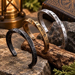 May include: Three open cuff bracelets in black, gold, and silver tones, each featuring a twisted metal design and engraved runic characters along the outer surface. The bracelets are arranged in a row, showcasing their metallic finishes and textures.
