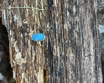 Op de afbeelding: Een ring met een ovale, levendig blauwe cabochonsteen in een eenvoudige metalen band. De steen vertoont subtiele interne kleurvariaties en een glad, gepolijst oppervlak, stevig vastgehouden in een dunne, metalen zetting.