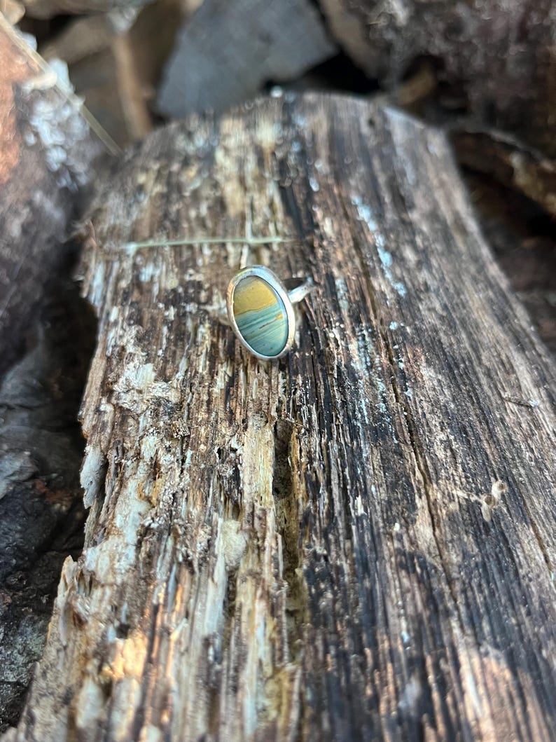 Op de afbeelding: Een zilveren ring met een ovaalvormige steen met horizontale banden in mosterdgeel, saliegroen en leiblauw. De steen is gezet in een gladde, gepolijste zilveren zetting en de ringband is gemaakt van een eenvoudige, gladde zilveren draad.