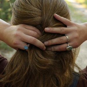 Op de afbeelding: Twee zilveren ringen met ovaalvormige stenen. De ene ring heeft een gladde, lichtblauwe steen, terwijl de andere ring een veelkleurige steen met tinten blauw, groen en bruin bevat. Beide ringen hebben dunne, eenvoudige metalen banden.