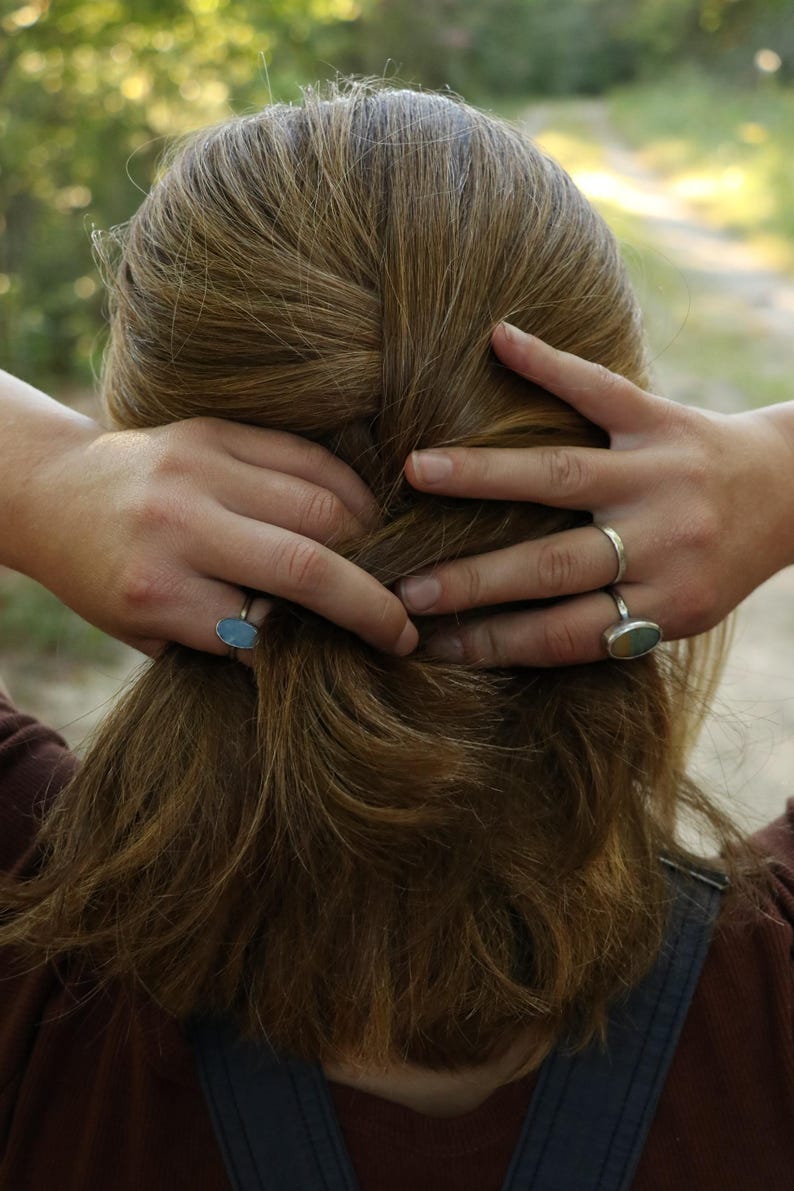 Op de afbeelding: Twee zilveren ringen met ovaalvormige stenen. De ene ring heeft een gladde, lichtblauwe steen, terwijl de andere ring een veelkleurige steen met tinten blauw, groen en bruin bevat. Beide ringen hebben dunne, eenvoudige metalen banden.
