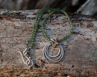 Op de afbeelding: Een ketting met een streng kleine, gefacetteerde groene kralen. De hanger bestaat uit drie concentrische zilverkleurige ringen die hangen aan een klein, rechthoekig messingkleurig metalen onderdeel. Een zilverkleurige haaksluiting is bevestigd.