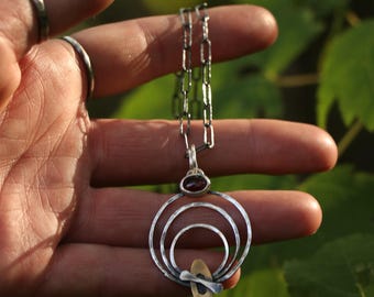 Op de afbeelding: Een zilveren hangerketting met drie concentrische gehamerde cirkels, een kleine donkerrode ovale edelsteen aan de bovenkant en een klein goudkleurig ovaal accent aan de onderkant, hangend aan een donkere geoxideerde zilveren schakelketting.