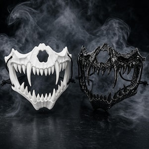 Peut inclure: Deux masques demi-visage présentant un design de mâchoire animale squelettique avec des crocs proéminents et des dents acérées. Un masque est blanc mat et l'autre est noir brillant. Les deux masques incluent des cordons élastiques noirs sur les côtés.