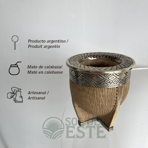 Pode incluir: Uma cuia de mate com exterior texturizado em castanho claro e uma borda de metal prateado com um padrão geométrico intrincado. A cuia possui três pés de metal prateado na base. O texto inclui Producto argentino, Mate de calabaza, Artesanal e SO DE ESTE.