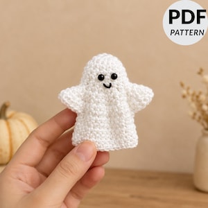 May include: A white crocheted ghost figure with two small black bead eyes and a stitched black mouth. The ghost has a rounded head and two outstretched arms. A circular white badge in the top right corner displays the text PDF PATTERN.