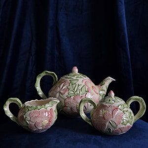 May include: A ceramic tea set comprising a teapot, a creamer, and a lidded sugar bowl. Each item displays a raised, embossed floral design in shades of pink and green on a cream-coloured base, complete with sculpted green handles and matching lids.