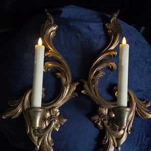 May include: A pair of gold-coloured wall sconces featuring ornate, swirling acanthus leaf carvings and floral accents at the base. Each sconce holds a single off-white, battery-operated taper candle with a glowing, flame-shaped LED tip.