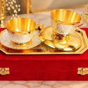 May include: Two metallic tea cups with matching saucers and a rectangular tray, all featuring a gold interior and a silver exterior with intricate floral engravings. The set is displayed on top of a rectangular red velvet box with gold latches.