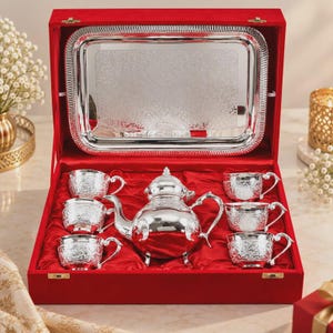 Puede incluir: Un juego de té de plata compuesto por una tetera y seis tazas a juego, todas con intrincados patrones florales en relieve. El conjunto se presenta en un estuche rectangular forrado de terciopelo rojo, que incluye una bandeja de plata.