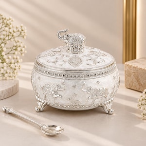 Puede incluir: Un recipiente redondo blanco y plateado con tapa, que presenta motivos de elefantes en relieve en el cuerpo y una figura de elefante esculpida en la tapa. El recipiente se apoya sobre cuatro patas ornamentadas y viene con una cuchara plateada a juego.