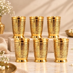 May include: A set of six gold-coloured metal tumblers arranged in two rows. Each tumbler features a detailed, intricate floral and vine pattern etched across the surface, with a smooth, polished rim and a sturdy, slightly flared base design.