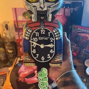 May include: A custom-painted Kit-Cat clock featuring a stylized character with a metallic mask, yellow eyes, and a red vest. The clock face is black with white numerals and the text Kit-Cat. A green, graffiti-style tail hangs from the bottom.
