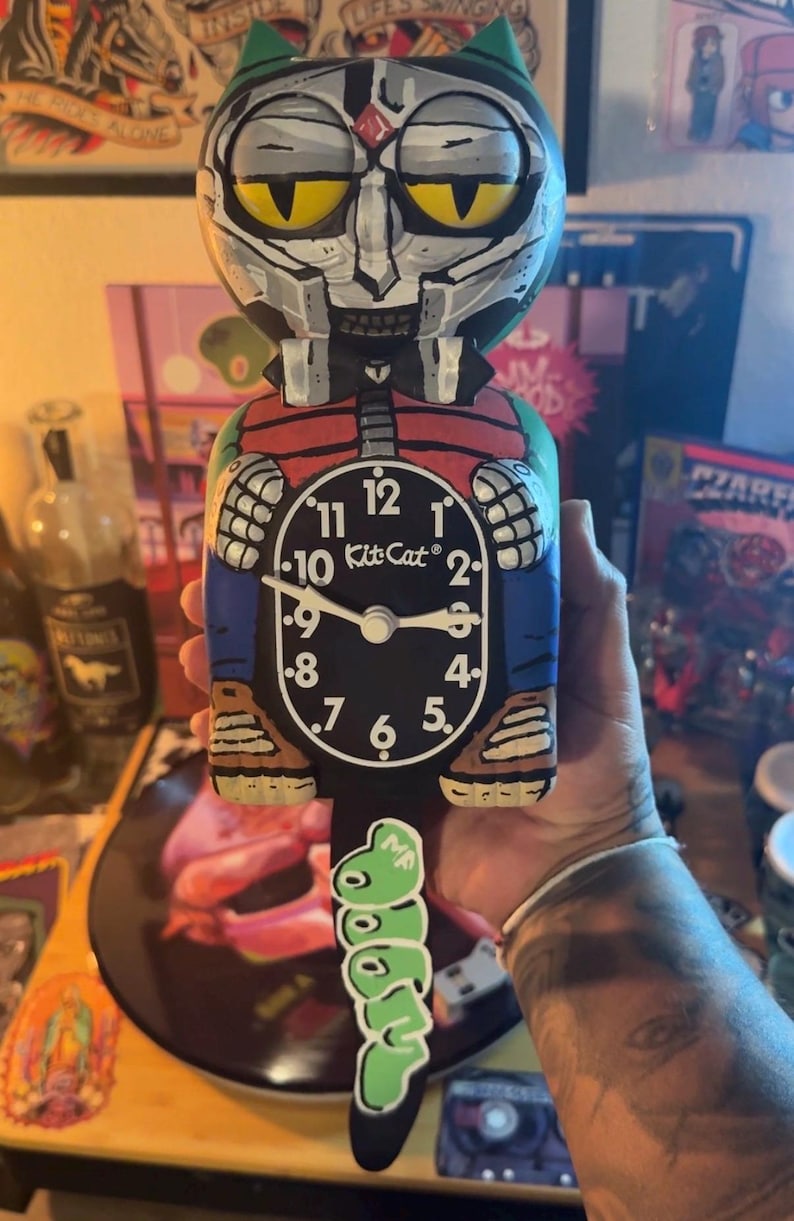 May include: A custom-painted Kit-Cat clock featuring a stylized character with a metallic mask, yellow eyes, and a red vest. The clock face is black with white numerals and the text Kit-Cat. A green, graffiti-style tail hangs from the bottom.