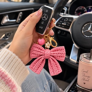 Puede incluir: Un llavero de coche Mercedes-Benz negro unido a una anilla de metal dorado con un gran lazo rosa de ganchillo texturizado, un pequeño dije de flor blanca con centro dorado y una cuenta rosa lisa, redonda y brillante.