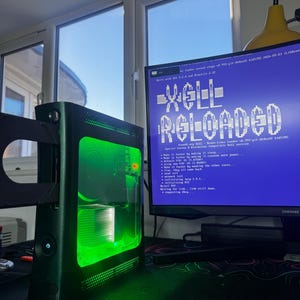 May include: A black games console with a glowing green interior and an open disc tray, connected to a computer monitor displaying the text Xell Reloaded in white letters on a blue background, showing system initialisation logs and technical data.