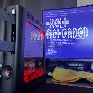May include: A black games console with a transparent side panel revealing internal red lighting and a metallic heatsink. The console is connected to a monitor displaying a blue screen with the white text XeLL RELOADED and system boot logs.