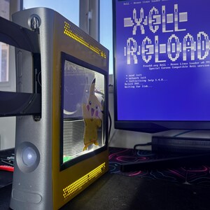 May include: A modified silver and yellow games console featuring a transparent side panel with a Pikachu decal, displaying internal components. Next to it, a computer monitor shows a blue screen with the text XeLL RELOAD and system logs.
