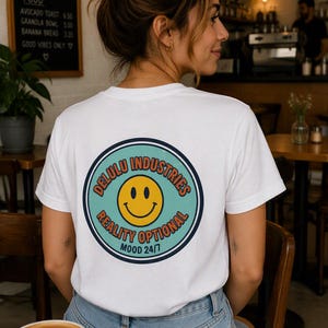 Könnte beinhalten: Ein weißes Kurzarm-T-Shirt mit einer kreisförmigen Grafik auf der Rückseite. Das Design zeigt ein gelbes Smiley-Gesicht, einen blaugrünen Hintergrund und dunkelblaue Ränder. Der Text lautet Delulu Industries, Reality Optional und Mood 24/7 in Orange.