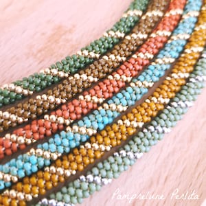 Può includere: Cinque collane di perline disposte in parallelo, ognuna caratterizzata da un motivo a spirale di perline colorate opache alternate a perline metalliche dorate o argentate. I colori includono verde oliva, marrone, terracotta, turchese e giallo senape.