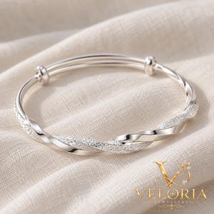 May include: A silver-toned bangle bracelet featuring a twisted design with alternating smooth polished sections and textured, glittery segments. The bracelet has an adjustable sliding mechanism with two small cylindrical beads at the closure.