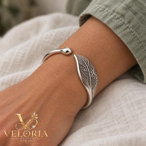Puede incluir: Una pulsera tipo brazalete de plata con un extremo redondeado y pulido y un terminal texturizado en forma de hoja. El diseño de la hoja incluye detalles intrincados de nervaduras oscuras. La pulsera es abierta.
