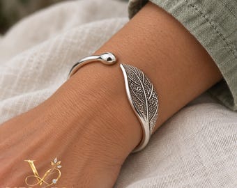 Op de afbeelding: Een zilveren manchetarmband met een gepolijst, afgerond uiteinde en een getextureerde, bladvormige terminal. Het bladontwerp bevat gedetailleerde, donkergekleurde nerven. De armband is aan één zijde open.