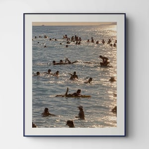 Könnte beinhalten: Ein gerahmter, vertikaler Wandkunstdruck, der eine sonnendurchflutete Meereslandschaft mit zahlreichen Menschen zeigt, die im Wasser schwimmen und surfen. Das Sonnenlicht erzeugt einen hellen, reflektierenden goldenen Glanz auf der Oberfläche des blauen Meeres, wobei Silhouetten von Personen über die Wellen verteilt sind.