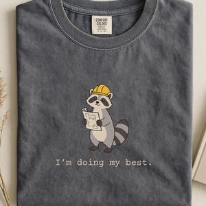 May include: A charcoal gray crew neck t-shirt featuring a graphic of a raccoon wearing a yellow hard hat and holding a blueprint. Below the raccoon, the text reads, I'm doing my best. The shirt has a Comfort Colors brand label at the inner neckline.